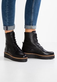 Botas de tobillo de cuero negro con cordones y suela de plataforma gruesa, con costuras en beige contrastantes, combinadas con jeans de denim con los puños enrollados.