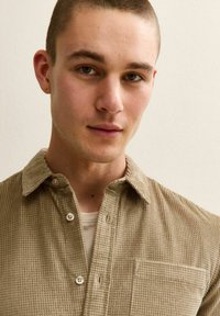 Jeune homme aux cheveux courts portant une chemise beige texturée boutonnée par-dessus un t-shirt blanc, regardant droit devant lui sur un fond uni.