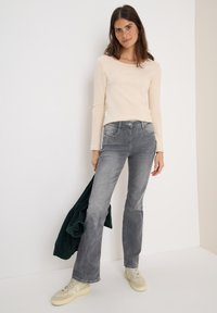 Top beige à manches longues, jean gris évasé avec poches, et baskets crème. Le mannequin tient une veste vert foncé, se tenant contre un fond neutre.
