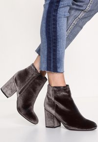 Botines de terciopelo gris oscuro con un alto tacón cuadrado y una cremallera lateral. La textura suave y la silueta elegante tienen un aspecto moderno y refinado.