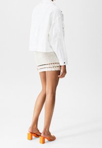 Biała krótka kurtka z długimi rękawami, kremowa mini spódnica szydełkowa z poziomymi wzorami oraz pomarańczowe sandały na blokowym obcasie.