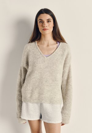 Pull en tricot surdimensionné gris clair avec un col en V et des poignets en côtes, porté par-dessus un débardeur lavande, associé à un short gris. Texture douce visible.