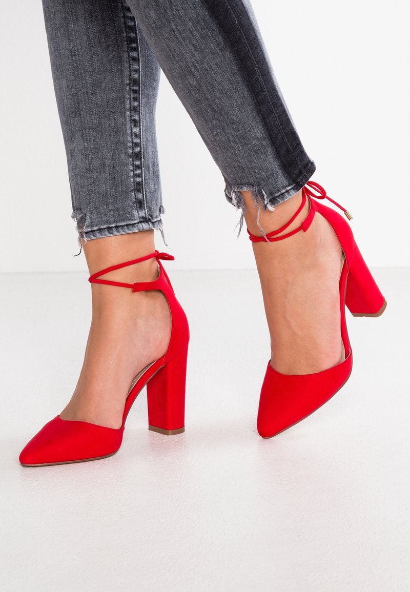Talons hauts en daim rouge avec un bout pointu, des brides à la cheville et un talon épais. Portés avec des jeans gris à ourlet effiloché.