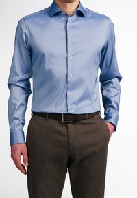Uomo che indossa una camicia elegante aderente a maniche lunghe di colore azzurro chiaro, infilata in pantaloni eleganti marroni con cintura nera, con la mano in tasca.
