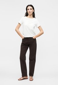 Femme debout, les mains dans les poches, portant une chemise blanche à manches courtes, un pantalon droit foncé et des sandales marron sur un fond uni.