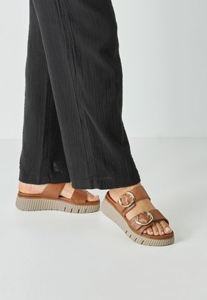 Bruine platformsandalen met twee brede banden met ronde metalen accenten en een beige, getextureerde zool. Gecombineerd met zwarte wijde broekspijpen.