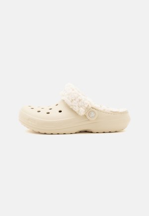 Beige slip-on træsko med ventilationshuller og hvid blød foring omkring hælkanten, vist fra siden på en ensfarvet baggrund.