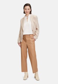 Blazer beige in tessuto testurizzato, camicia bianca con cravatta al collo e pantaloni in pelle beige. Modello con stivaletti crema alla caviglia.