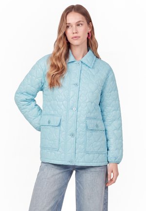 Femme portant une veste matelassée bleu clair avec des motifs en forme de cœur, fermeture à boutons, poches à rabat et jean bleu clair, debout avec une main sur la hanche.