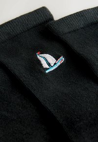 Chaussettes noires ornées d'un petit voilier brodé en blanc, bleu et rouge. Le tissu est lisse avec des bords côtes aux chevilles.