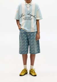 Světle modrá a krémová pruhovaná polokošile s krátkým rukávem a nápisem, doplněná světle džínovými šortkami s opakujícím se logem. Žluté tenisky.