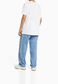 Person som bär en enkel vit t-shirt, ljusblå baggy jeans med sömdetaljer och vita sneakers, står vänd bort mot en vit bakgrund.