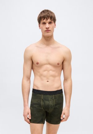 Jeune homme aux cheveux bruns courts portant un boxer vert foncé camouflage avec une ceinture noire, debout devant un fond gris clair uni.