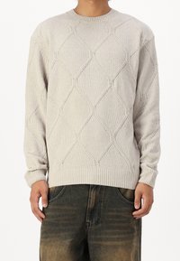 Homme portant un pull en tricot beige clair à motif losange et un jean foncé délavé, debout devant un fond blanc.