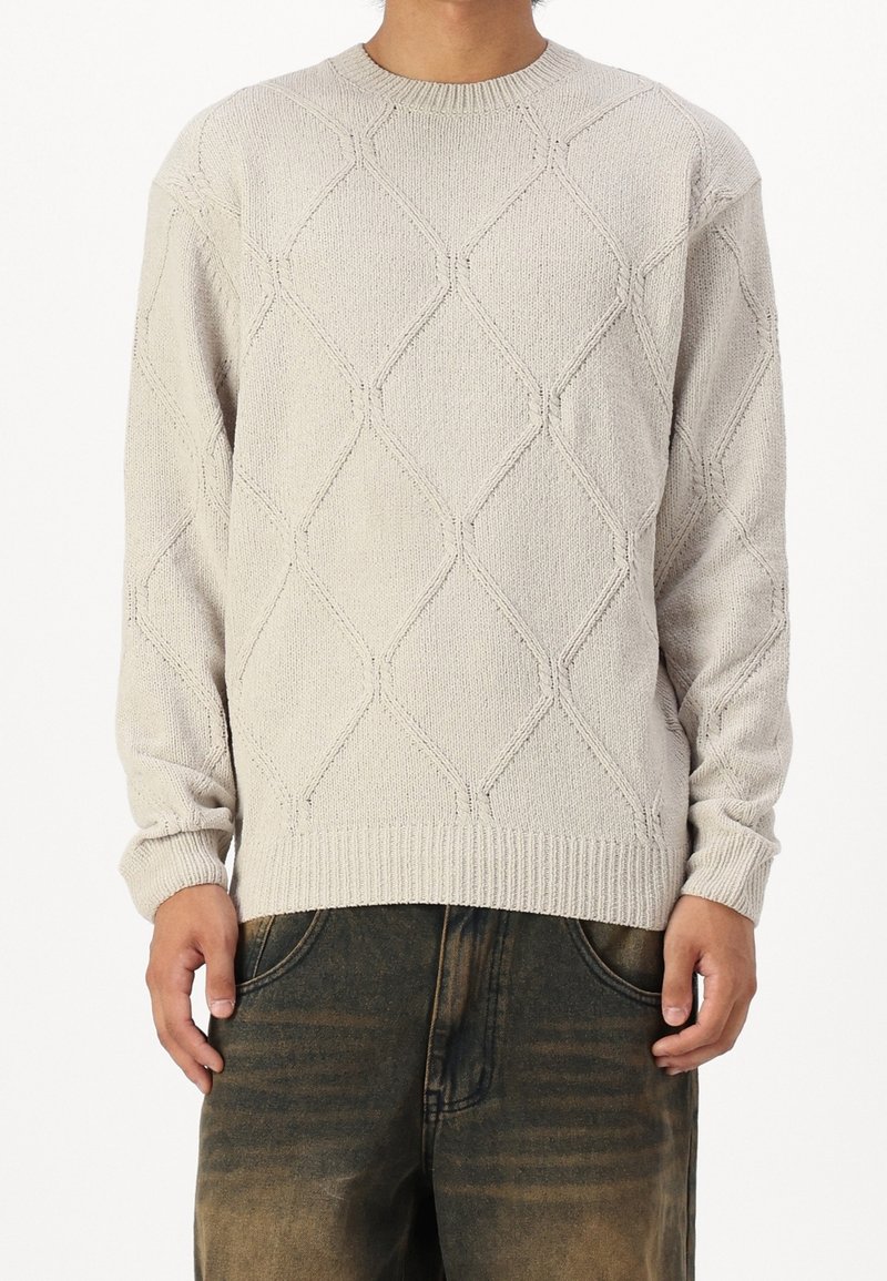 Homme portant un pull en tricot beige clair à motif losange et un jean foncé délavé, debout devant un fond blanc.