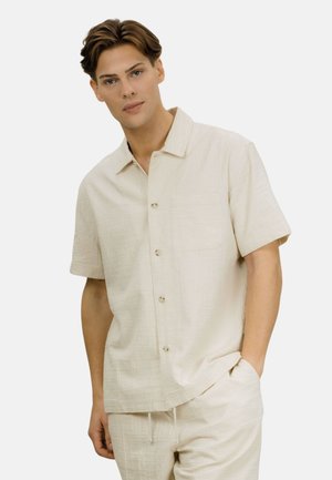 Ung mann iført en kremfarget kortermet skjorte med knapper og matchende bukser, stående med én hånd i lomma mot en ensfarget bakgrunn.