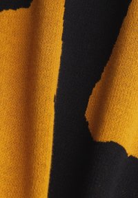 Stickad tyg med kraftiga gula och mörkblå sektioner samt en texturerad yta. Mönstret framstår som abstrakt och lager på lager.