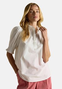 Frau mit langen blonden Haaren, die eine weiße Bluse mit Puffärmeln und rosa Cordhosen trägt, posiert mit einer Hand nahe ihrem Gesicht.