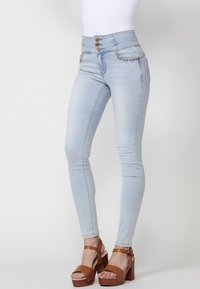 Jeans ajustados de tiro alto en azul claro con cierre de tres botones, detalle de cadena en los bolsillos y una textura de mezclilla suave. Sandalias de plataforma marrones.