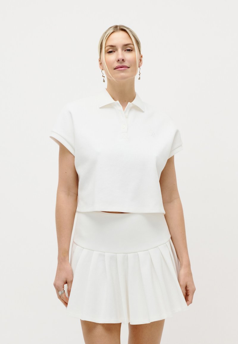 Ξανθιά γυναίκα με άσπρο κοντό πόλο πουκάμισο και πλισέ φούστα, που στέκεται μπροστά σε απλό λευκό φόντο.