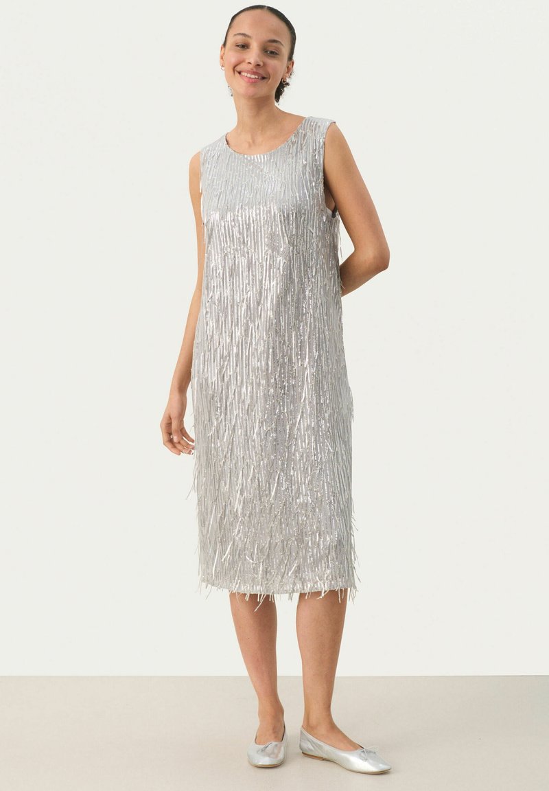 Vestido plateado sin mangas con detalles de flecos verticales, hasta la rodilla, de textura suave, combinado con zapatos planos plateados. Diseño sencillo y elegante.