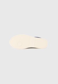 Semelle de chaussure en caoutchouc, présentant une texture lisse beige avec un emblème circulaire central et un motif antidérapant pour l'adhérence.