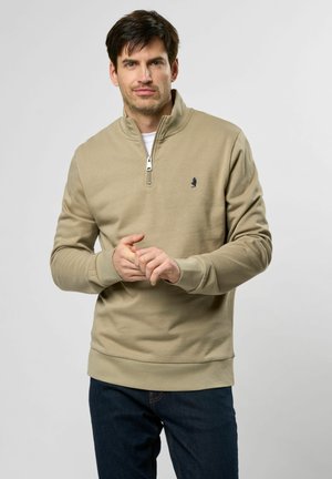Beige Sweatshirt mit Reißverschlusskragen und gerippten Bündchen, mit einem kleinen blauen Logo auf der Brust. Getragen mit dunkelblauen Hosen.