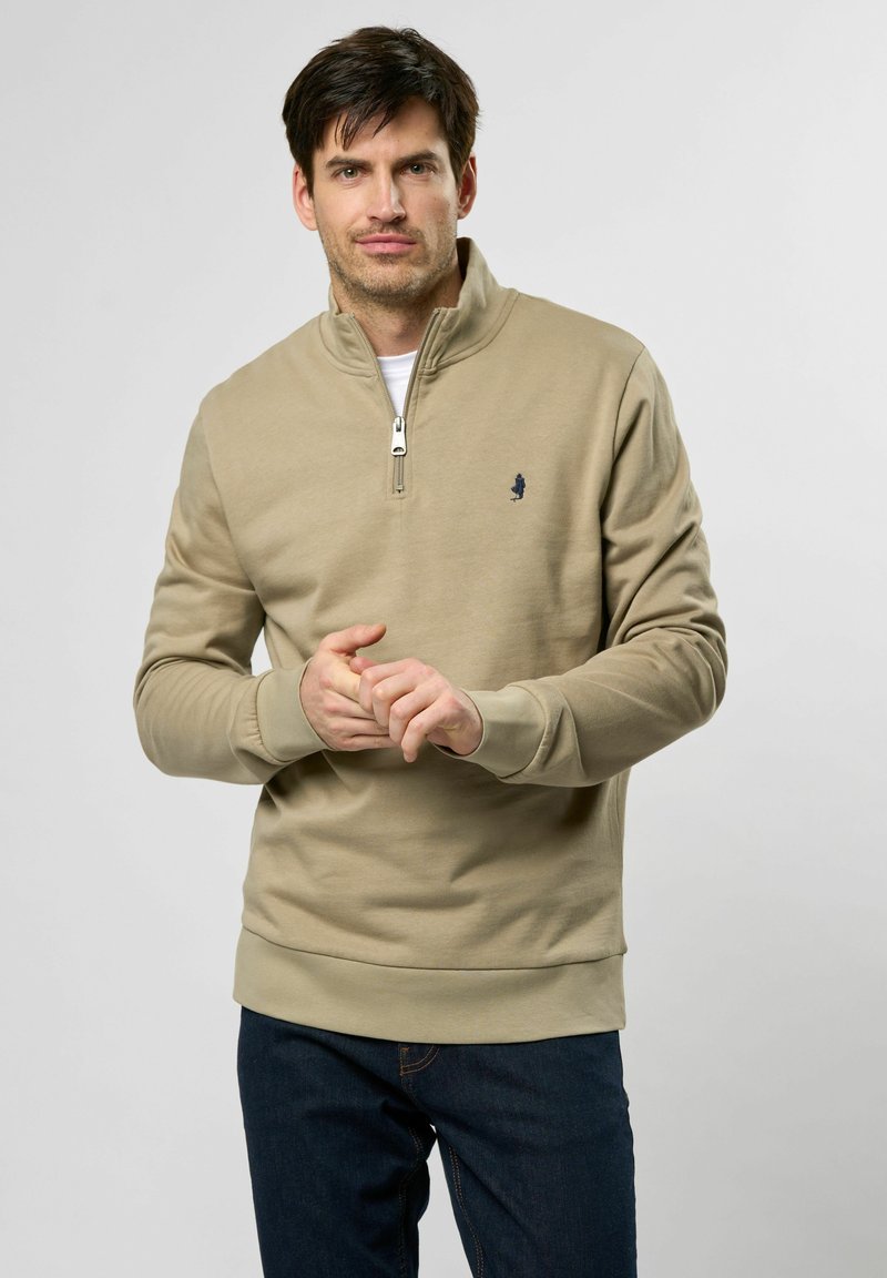 Beige genser med glidelås i halsen og ribbestrikkede mansjetter, med en liten marineblå logo på brystet. Brukes med mørkeblå bukser.