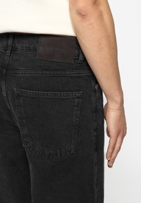 Κοντινή λήψη της μέσης ενός ατόμου που φορά μαύρο τζιν παντελόνι με πίσω τσέπη και σκούρο ετικέτα επωνυμίας στη ζώνη.