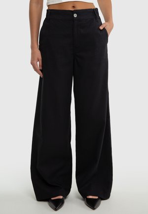 Mujer con pantalones negros de pierna ancha y cintura alta, con botones delanteros y bolsillos, combinados con zapatos negros de punta afilada y un top corto blanco.