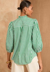 Blusa a righe verdi e bianche con scollo arricciato, maniche a sbuffo e orlo curvo. Realizzata in tessuto leggero.
