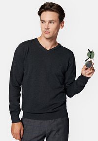 Ciemnoszary sweter w serek z miękkiego dzianiny, z prążkowanymi mankietami i dołem. Model trzyma okrągłe okulary przeciwsłoneczne w zielonym odcieniu.