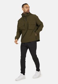 Olijfgroen pullover jack met twee grote voorzakken, een halverwege ritssluiting en elastische manchetten, gecombineerd met zwarte slim-fit broeken en witte sneakers.