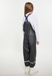 Juodos vandeniui atsparios kombinezonas su elastingu liemeniu, reguliuojamomis mėlynomis petnešomis ir baltais atspindinčiais dryžiais ant rankovių, derintas su baltais batais.