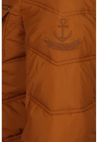 Brun quiltet jakke stof med en broderet anker-emblem over en delvist synlig lomme med en metallisk knap.