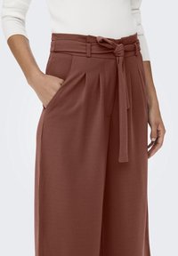 Pantalon à jambes larges de couleur rouille, doté d'une ceinture à la taille, de plis à l'avant et de poches latérales. Tissu doux avec une texture lisse.