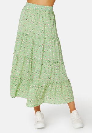 Vrouw draagt een groene midi-rok met bloemenprint en meerdere lagen, gecombineerd met witte vetersneakers, staand tegen een witte achtergrond.