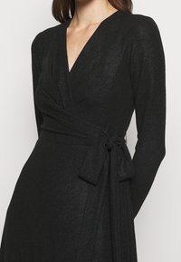 Nainen, jolla on päällään teksturoitu musta kietaisumekko pitkähihaisilla hihoilla ja vyötärölle solmittu vyö yksinkertaista taustaa vasten.