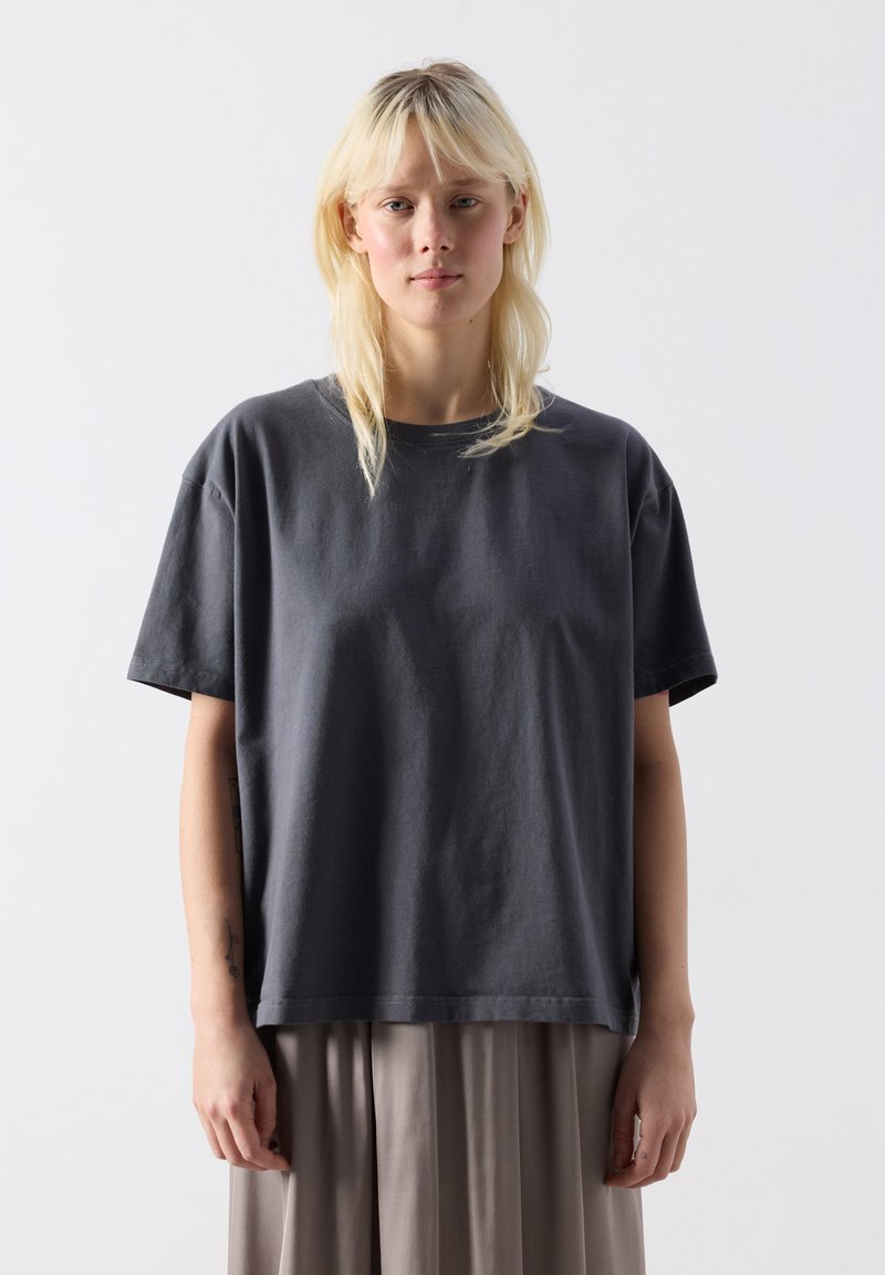 Mladá osoba so svetlými vlasmi, oblečená v voľnom tmavosivom tričku a svetlosivých plisovaných nohaviciach, stojaca pred jednoduchým bielym pozadím.