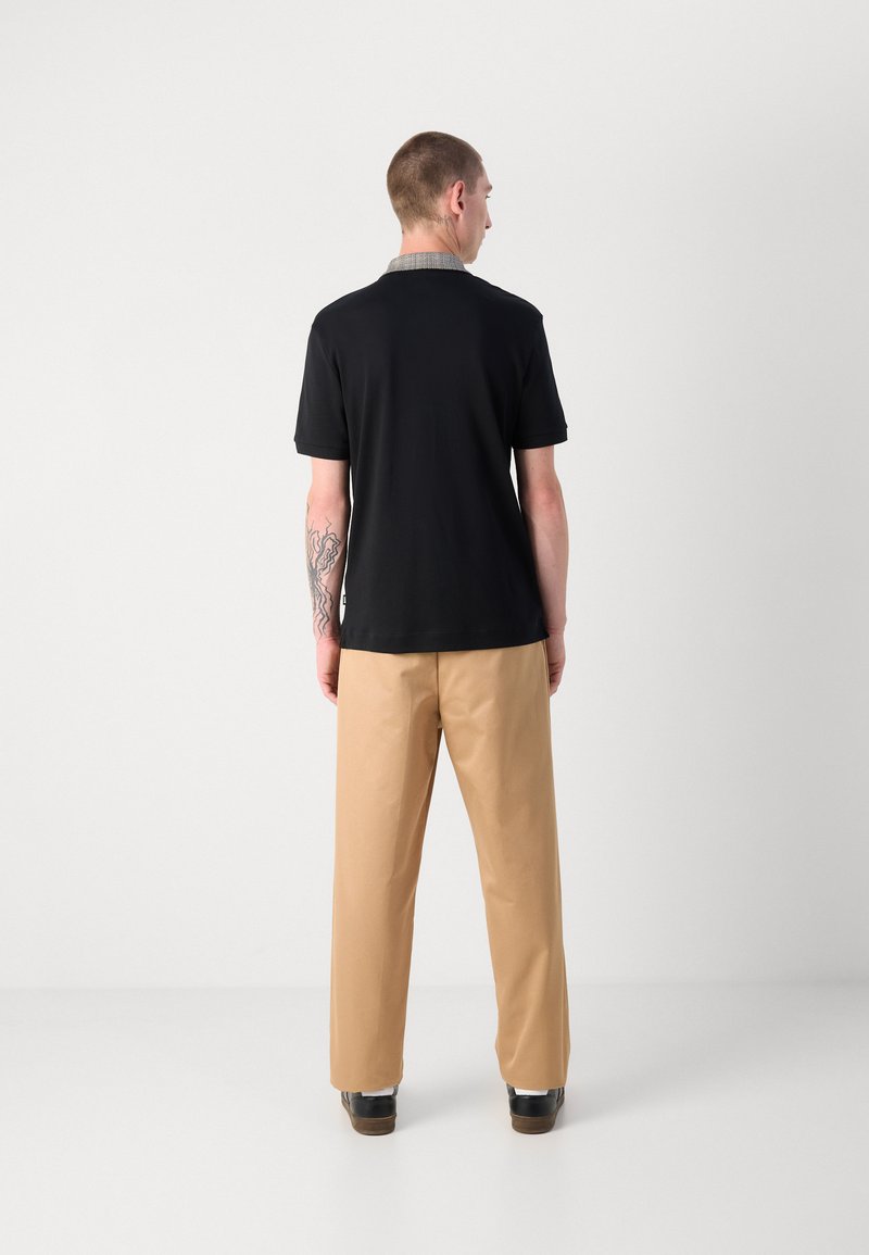 Schwarzes Poloshirt mit einem gemusterten Kragen, kombiniert mit beigen Hosen. Der Stoff scheint glatt zu sein, mit lockerer Passform und kurzen Ärmeln.