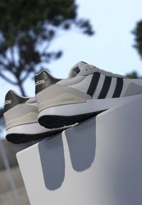 Paire de baskets Adidas blanches et noires posées sur un rebord blanc avec des arbres et un ciel flous en arrière-plan.