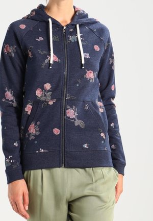 Marineblauer Kapuzenpullover mit Reißverschluss und pinkem Blumenmuster, weißen Kordeln, Vordertaschen, getragen mit hellgrüner Hose.