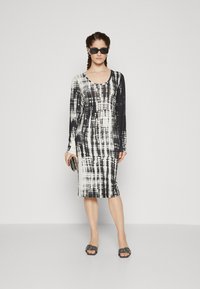 Long-sleeve, knee-length dress featuring a black and white tie-dye pattern, made from a smooth fabric. Model wears black sunglasses and sandals.