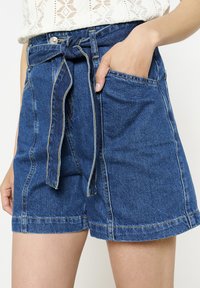 Denim šorti ar jostu viduklī, gaiši zils tonis, ar priekšējiem kabatām un kontrastējošām šuvēm, demonstrējot ikdienišķu un brīvu dizainu.