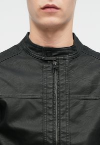 Giacca di pelle nera con colletto alto, superficie texture, zip centrale e dettagli di cucitura orizzontale sulle spalle.