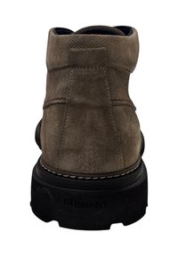 Bruine suède enkelboot met een gestructureerde bovenkant, versterkte stiksels en een zwarte rubberen zool met de merknaam "Fluchos" in reliëf.