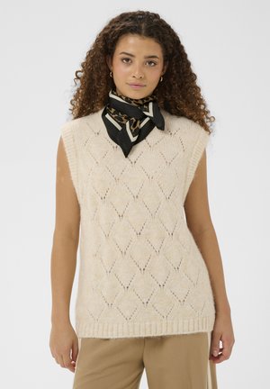 Ženska s kodrastimi lasmi nosi bež brezrokavnik iz pletenine, črno šal z leopardjim vzorcem, ki je zavezana okoli vratu, in barvne hlače na enostavni podlagi.