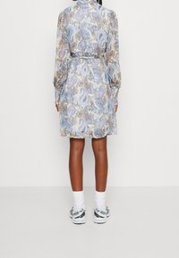Robe à imprimé paisley floral en bleu clair, beige et marron, avec des manches longues, une ceinture à la taille et une coupe décontractée. Des baskets sont portées.