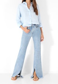 Chemise boutonnée bleu clair associée à un jean en denim clair à pattes d'éléphant avec des fentes latérales et des ourlets effilochés. Des sandales beiges complètent l'ensemble.