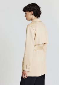 Donna con capelli ricci corti, vestita con un trench coat beige con cintura e pantaloni neri, rivolta di lato contro uno sfondo semplice.